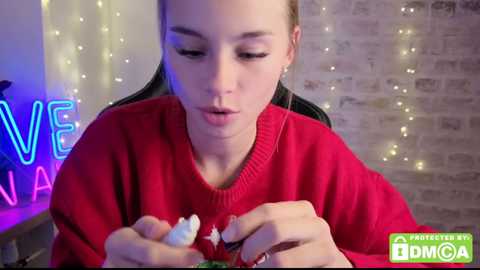 Media: Video of a young woman with light skin and straight, light brown hair, wearing a red sweater, intently crafting something with her hands. Background features a brick wall and a glowing \"V\" neon sign.