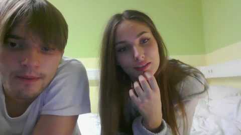 Video of a young couple: a light-skinned, brunette girl with long hair, wearing a gray shirt, and a light-skinned, short-haired boy in a white shirt, both looking at the camera in a brightly lit room with green walls and white furniture.