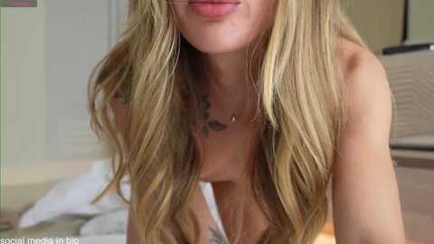 Video of a blonde woman with long, wavy hair, wearing a white top, leaning forward with her breasts slightly exposed. Background shows a bed and window blinds.