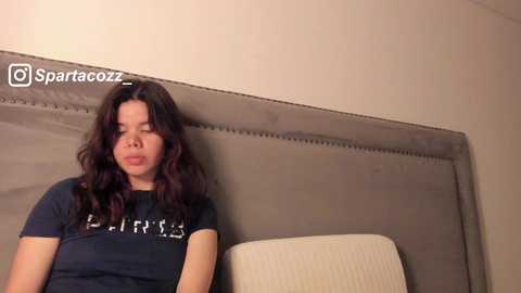 Media: Video of a young woman with long, wavy dark hair, wearing a black T-shirt with white text, leaning against a gray upholstered headboard, looking pensive.