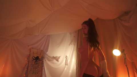 Video of a woman in a tent, holding a white sheet. She has long dark hair and is wearing a pink jacket. The background is dimly lit, with a warm orange glow.