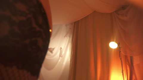Video of a dimly lit room with a warm, orange glow. A large, soft light fixture casts a circular light on the ceiling, while the walls are draped in sheer, white curtains. The ambiance is cozy and intimate.