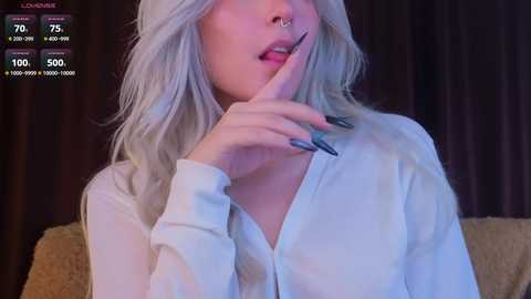 Video of a fair-skinned woman with platinum blonde hair, wearing a white button-up shirt, holding a finger to her lips. Background shows dim lighting and a beige couch.