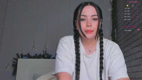 Video of a young woman with long black braids, fair skin, and a white t-shirt, sitting against a grey brick wall adorned with fairy lights.