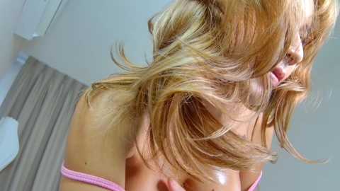 Video of a blonde woman with wavy hair, wearing a pink off-shoulder top, leaning forward, with a blurred background featuring white curtains and a beige wall.