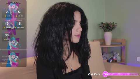 Video of a young woman with long, black hair, wearing a black top, in a green room with a flower vase and a bookshelf.
