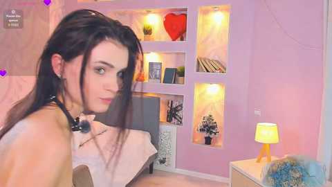 Media: Video of a young woman with long black hair, wearing a white top, standing in a pink room with a bookshelf, heart-shaped red light, and a blue-patterned chair.