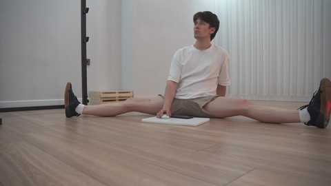 Video of a young Caucasian man with curly hair in a white T-shirt and shorts, performing a split on a wooden floor, in a sparsely furnished room with a tall black pole and a box in the background.