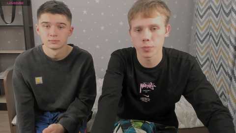 Video of two young men, one with short black hair in a gray sweatshirt, the other with short red hair in a black shirt, sitting on a bed with a gray patterned wall and a zigzag-patterned curtain in the background.