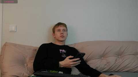 Video of a young man with short, light brown hair, wearing a black long-sleeve shirt, seated on a light pink, tufted sofa against a plain white wall, holding a black smartphone.