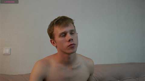Video of a shirtless, fair-skinned, young man with short, light brown hair, sitting on a beige couch against a plain, light gray wall. He appears thoughtful.