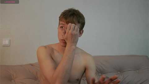 Video of a shirtless young Caucasian man with short brown hair, covering his face with one hand, sitting on a light gray tufted couch against a light blue wall, looking introspective.