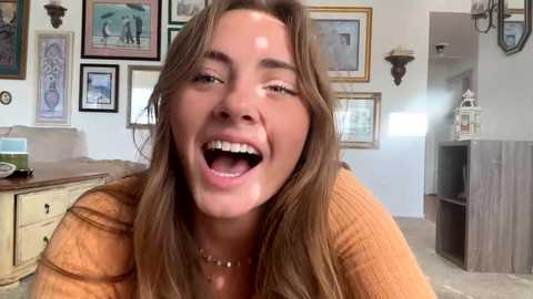 Media: Video of a smiling, light-skinned woman with long brown hair, wearing a mustard-yellow sweater, in a cozy living room adorned with framed art and a rustic wooden shelf.