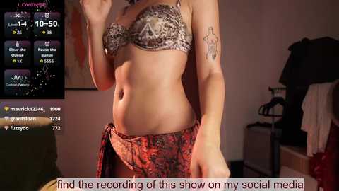 A video of a woman with a tattooed arm, wearing leopard print lingerie and red pants, taken in a dimly lit bedroom. The background shows a cluttered room with a TV displaying a chat feed.
