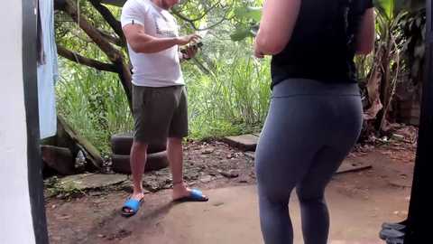 Video of a muscular man in a white t-shirt and shorts, showing off a blue flip-flop, interacting with a woman in a black tank top and grey leggings, standing in a lush, green, outdoor setting.