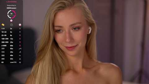 A video of a topless blonde woman with long, straight hair, fair skin, and a slender physique. She has a soft smile. The background is blurred, featuring a purple curtain.