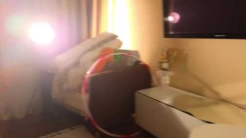 A video of a cluttered hotel room with a blurry, bright light source, a bed with a red pillow, a TV, and a glass of water on a nightstand.