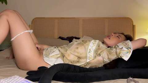 A video of an Asian woman lying on a bed, wearing a floral shirt and white panties, legs spread, revealing a penis, in a dimly lit room.
