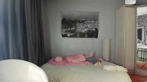 Video of a serene, light-filled bedroom with a gray wall, a black-and-white cityscape print, a white bed with pink pillows, and a white chair.