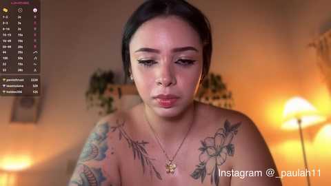 A video of a tattooed woman with medium skin tone and black hair, wearing a gold necklace, in a dimly lit room with potted plants and a lamp.