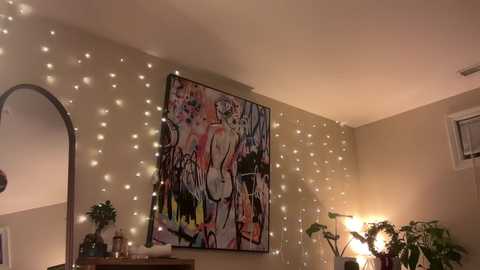 Video of a warmly lit bedroom with beige walls adorned with a large, colorful abstract painting, fairy lights, and potted plants.