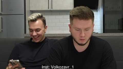Video of two men in a modern kitchen; one with short, blonde hair, the other with short, brown hair, both smiling, wearing black t-shirts. White subway tiles in the background.