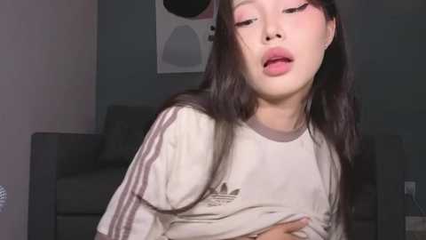 Video of an Asian woman with long black hair, wearing a white Adidas shirt, lying on a couch, eyes closed, lips parted, in a dimly lit room.