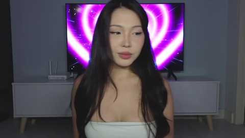 Media: Video of a young Asian woman with long black hair, wearing a white strapless top, in a modern living room with a purple and pink circular light fixture on the wall and white furniture.