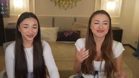 Video of two smiling women with long, straight brown hair, wearing white tops, sitting on a couch in a modern, beige bedroom with a large bed, wall art, and lamps.