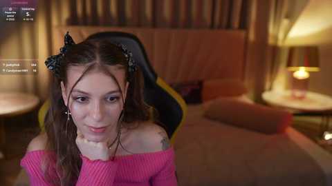 Media: A young woman with long brown hair in pigtails, wearing a pink off-shoulder sweater, sits on a gaming chair in a dimly lit bedroom.