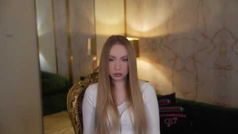 Video of a blonde woman with long hair, wearing a white top, seated on a green velvet couch in a dimly lit room with beige marble walls and a lit lamp.
