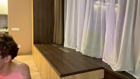 A video of a woman with short dark hair, wearing a pink tank top, standing beside a window with sheer white curtains, wooden paneling, and a light-colored wooden floor.