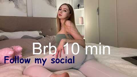 Video of a young, slender, fair-skinned woman with long blonde hair, wearing a strapless top and light-colored leggings, reclining on a bed in a modern, beige-toned bedroom. Text overlay reads: \"Bra 10 min show my social.\