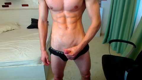 Media: Video of a lean, fit, light-skinned man in black briefs, standing in a modern bedroom with white bed, green curtains, and black chair.