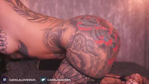 Video of a tattooed, light-skinned woman with a large, detailed back tattoo, wearing black fishnet stockings, kneeling on a dark carpet.