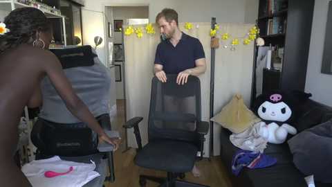 Media: Video of a bare-chested Black woman adjusting a chair, a bearded white man sitting, and a stuffed cat on a couch in a cluttered, brightly-lit living room.