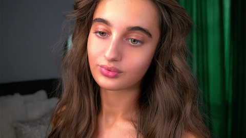 Video of a young woman with long, wavy brown hair, fair skin, and a neutral expression, wearing pink lipstick, standing against a dark background with a hint of green curtains.