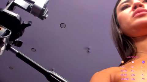 Video of a young woman with straight dark hair, wearing a black top, standing in front of a purple backdrop with a camera rig. The image includes a live stream overlay.