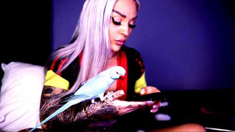 Video of a tattooed woman with long, platinum blonde hair and bold makeup, holding a small, light blue bird in a dimly lit room.