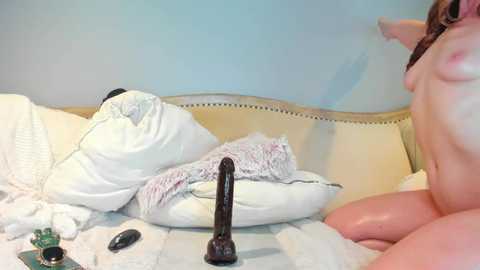 A video shows a naked woman with a large, black dildo, lying on a bed with white pillows and a beige headboard.