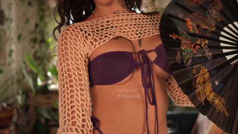 Video of a woman with light brown skin, wearing a crochet top and a purple bikini. She holds a fan with colorful designs. Background features greenery and wooden furniture.