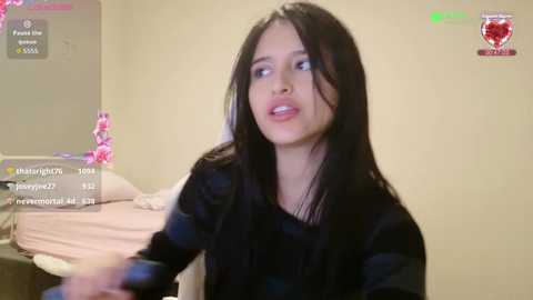 Video of a young woman with long black hair, wearing a black top, in a beige room with a bed and pink flowers, live-streaming on Twitch.
