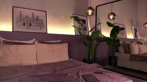 Media: Video of a modern, cozy bedroom with lavender walls, a large bed with beige linens, two potted plants, two pendant lights, and framed artwork.