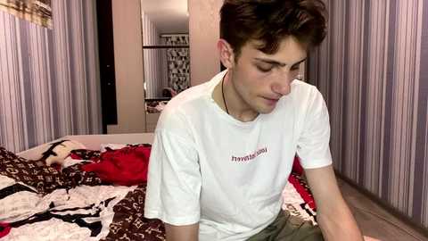 Video of a young man with short brown hair, wearing a white t-shirt, sitting on a bed with patterned blankets. Background shows a modern, minimalistic bedroom with striped wallpaper.