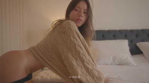 Video of a young woman with long brown hair, wearing a knit sweater, leaning over a bed, revealing her buttocks in a bedroom with a tufted headboard.