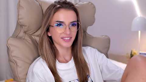 Video of a young Caucasian woman with long, straight brown hair, wearing clear glasses and a white shirt, sitting in a beige leather chair, smiling in a modern office setting.