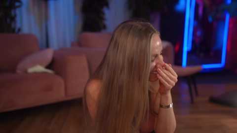 Video of a young woman with long, straight blonde hair, wearing a watch, kneeling on a wooden floor in a dimly lit living room with a pink sofa and colorful neon lights in the background.