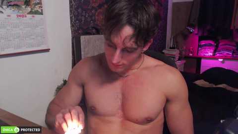 Video of a shirtless, muscular young man with short brown hair, holding a lit cigarette, in a dimly lit room with a calendar, shelves, and a purple light.