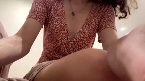 Video of a woman in a pink floral dress, leaning forward, showing cleavage, with a blurred background.