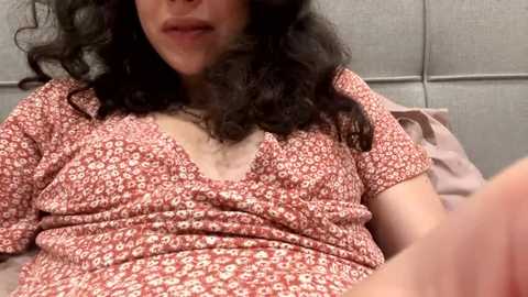 A video of a woman with curly dark hair, wearing a red floral dress, reclining on a bed with a grey headboard, in a cozy, intimate setting.